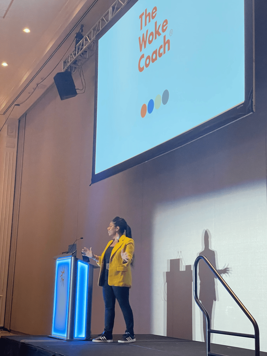 Seena Hodges, a Black woman in a yellow blazer, jeans and sneakers, speaks in front of a crowd at a podium at the Women in Agile conference. The screen behind her includes The Woke Coach in red.