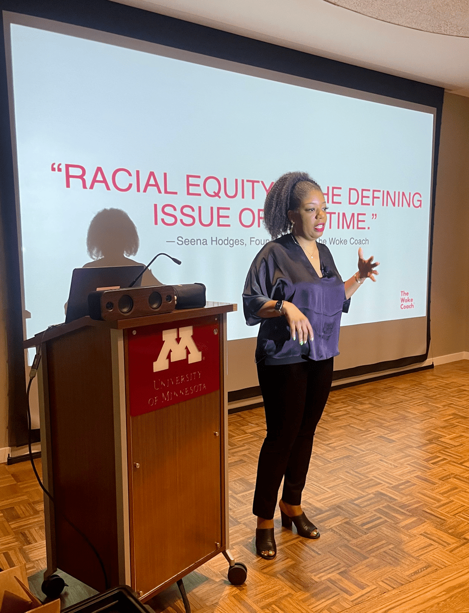 Seena Hodges, a Black woman in a purple top, black pants, and black heels, speaks to the University of Minnesota in front of a screen that reads "Racial equity is the defining issue of our time."